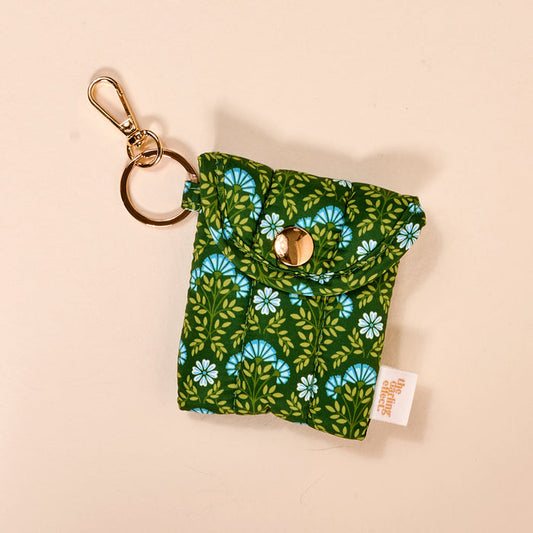 The Darling Effect's Everbloom Forest Green Puffy Earbud Case features green fabric with a blue and white floral print, gold snap closure, and a gold keychain clip—a stylish way to carry wireless earbuds. A white side tag is sewn into the seam.