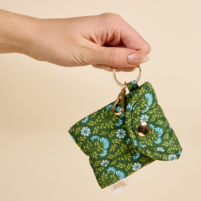 Hand holding The Darling Effect Everbloom Forest Green Puffy Earbud Case, featuring a quilted design with green floral pattern, gold snap closure, and keychain clip—ideal for wireless earbuds—against a beige background.