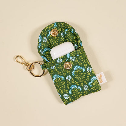 The Darling Effect Everbloom Forest Green Puffy Earbud Case holds white wireless earbuds. It features a floral design, gold keychain, snap button, small side tag, and is shown on a beige background.