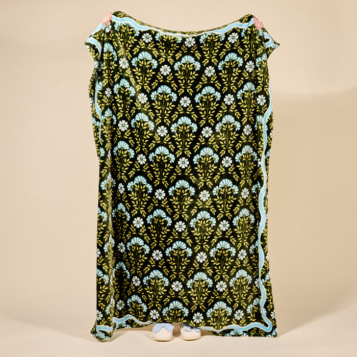 A person holds up The Darling Effect’s Everbloom Forest Green Snuggle Up Plush Blanket featuring a repeating green and blue floral pattern with a light blue border; only their hands and shoes are visible.