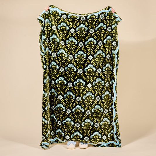 A person holds up The Darling Effect’s Everbloom Forest Green Snuggle Up Plush Blanket featuring a repeating green and blue floral pattern with a light blue border; only their hands and shoes are visible.