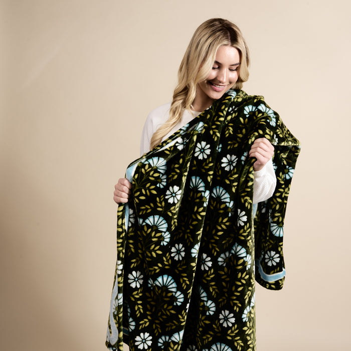 A woman with long blonde hair smiles while holding The Darling Effect's Everbloom Forest Green Snuggle Up Plush Blanket, featuring blue and white floral designs, against a beige background.