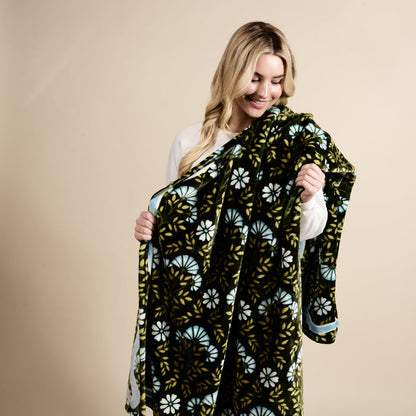 A woman with long blonde hair smiles while holding The Darling Effect's Everbloom Forest Green Snuggle Up Plush Blanket, featuring blue and white floral designs, against a beige background.