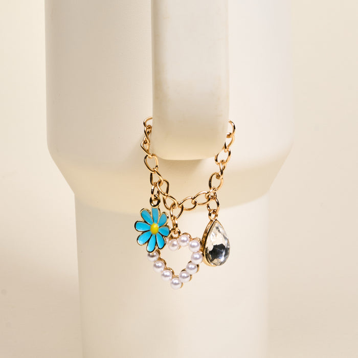 The Darling Effect's Fairytale Tumbler Charm, a gold chain bracelet with a blue enamel daisy, pearl heart outline, and teardrop crystal, is shown on a cream mannequin wrist—perfect for pairing with your favorite tumbler accessory.