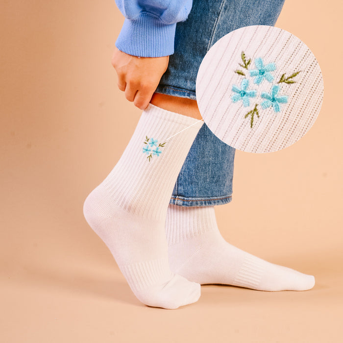 A person in rolled-up blue jeans and a blue sweater adjusts their white The Darling Effect Floral Blue Embroidered Crew Socks, featuring a small blue flower. A circular inset displays a close-up of the stylish embroidery on these women's socks.