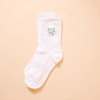 The Darling Effect’s Floral Blue Embroidered Crew Socks feature white ribbed fabric with a blue floral embroidery and green leaves—stylish socks that add a charming touch to any collection.
