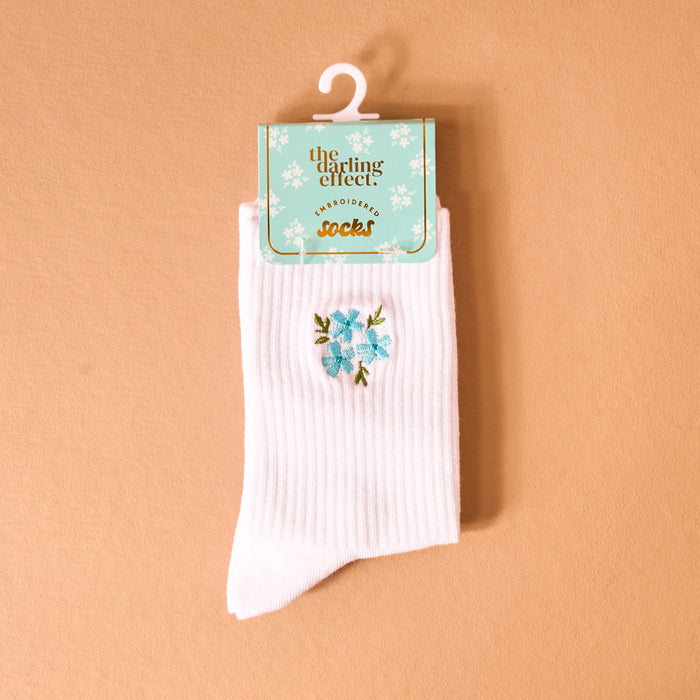 The Darling Effect Floral Blue Embroidered Crew Socks are shown in white with blue floral embroidery on a peach background, packaged with a teal tag and gold “EMBROIDERED socks” lettering.