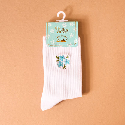 The Darling Effect Floral Blue Embroidered Crew Socks are shown in white with blue floral embroidery on a peach background, packaged with a teal tag and gold “EMBROIDERED socks” lettering.
