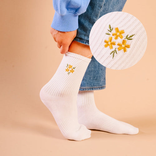 A person adjusts The Darling Effect’s Floral Yellow Embroidered Crew Socks, featuring colorful flower designs. A circular inset highlights the embroidery. Shown with blue jeans and a light blue sweater. Available in women’s sizes 5–10.
