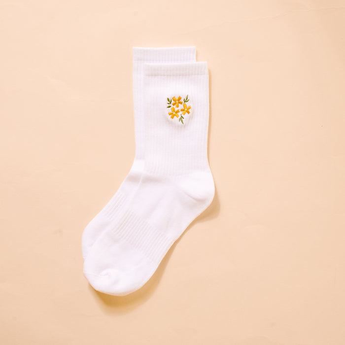 The Darling Effect Floral Yellow Embroidered Crew Socks feature a small yellow flower with green leaves on white fabric, shown on a light beige background. Available in women's sizes 5–10.