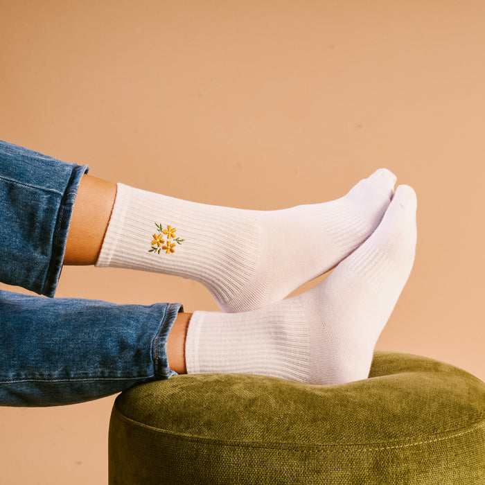 A person wearing The Darling Effect Floral Yellow Embroidered Crew Socks rests their feet on a green ottoman against a beige backdrop. Available in women's sizes 5–10, these socks feature charming, colorful floral embroidery.