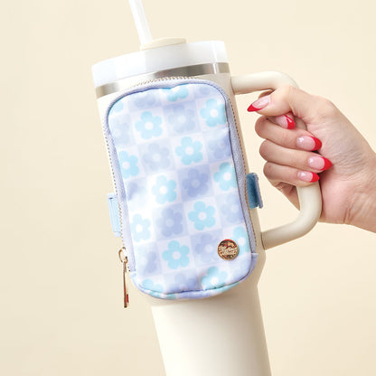 A hand with pink manicured nails holds a large white tumbler featuring The Darling Effect's Flower Check Blue Tumbler Fanny Pack, a water-resistant accessory in a blue checkered and floral pattern. The soft beige background adds warmth.
