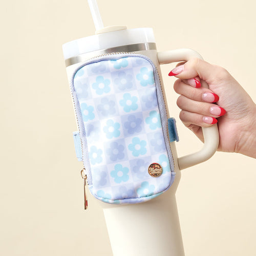 A hand with pink manicured nails holds a large white tumbler featuring The Darling Effect's Flower Check Blue Tumbler Fanny Pack, a water-resistant accessory in a blue checkered and floral pattern. The soft beige background adds warmth.