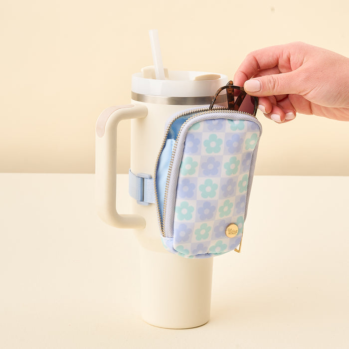 A hand places sunglasses into The Darling Effect’s Flower Check Blue Tumbler Fanny Pack, attached to the handle of a large cream-colored travel mug with a straw.