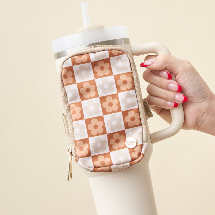 A hand with pink and white manicured nails holds a cream tumbler and The Darling Effect's Flower Check Brown Tumbler Fanny Pack, featuring a peach and beige checkered pattern with white and orange flowers.