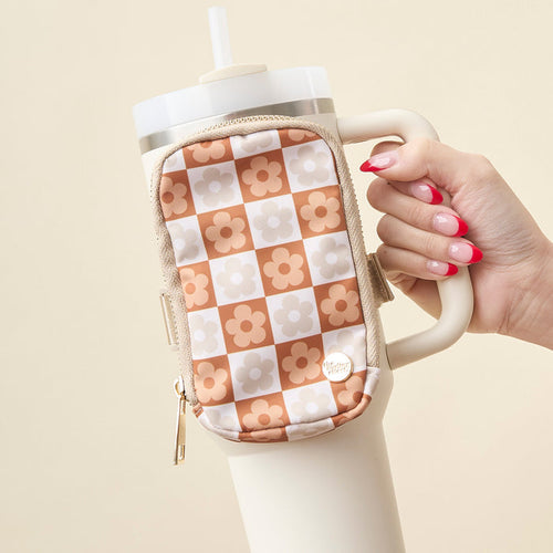 A hand with pink and white manicured nails holds a cream tumbler and The Darling Effect's Flower Check Brown Tumbler Fanny Pack, featuring a peach and beige checkered pattern with white and orange flowers.