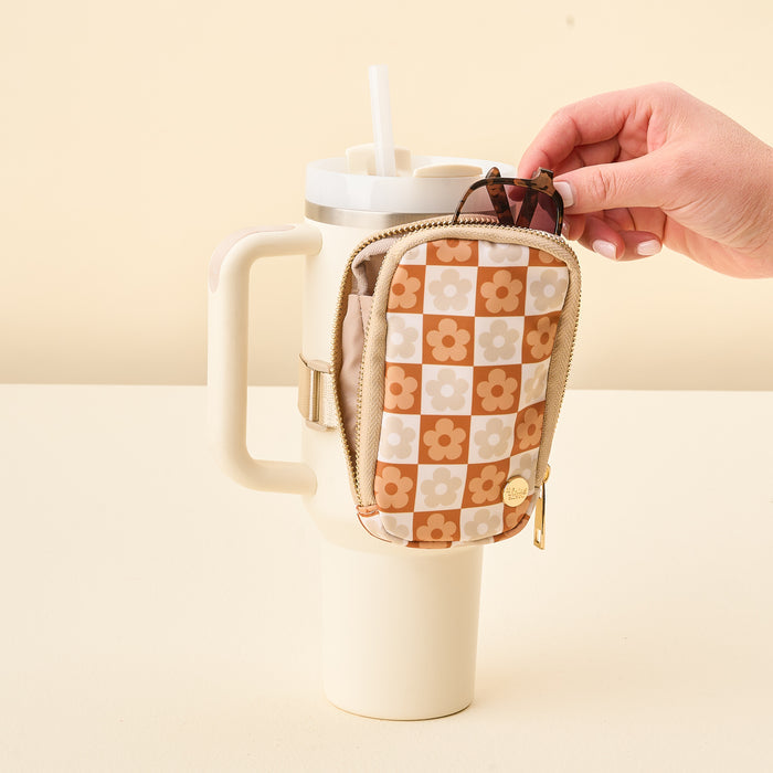 A hand attaches The Darling Effect Flower Check Brown Tumbler Fanny Pack, featuring a tan and orange floral checkerboard design, to the handle of a large cream tumbler with a straw.