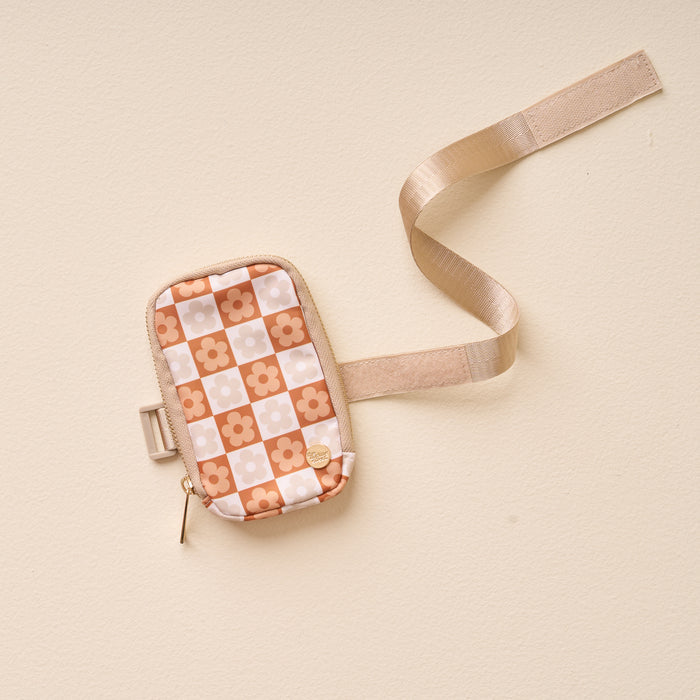 The Darling Effect’s Flower Check Brown Tumbler Fanny Pack, with a beige strap and retro orange and white checkerboard floral pattern, is displayed on a light beige surface.