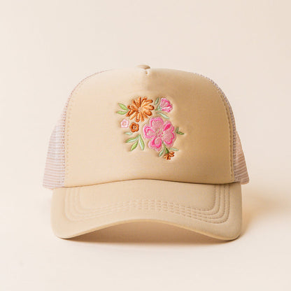 The Flowers Trucker Hat by the-darling-effect is a beige hat with mesh sides, showcasing an embroidered floral design of pink, orange, and yellow flowers with green leaves on the front.