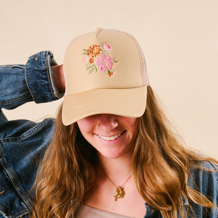 A smiling woman wears the-darling-effect’s Flowers Trucker Hat in cream with a pink and orange floral bouquet, styled with a denim jacket and gold necklace.