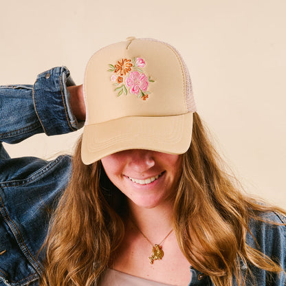 A smiling woman wears the-darling-effect’s Flowers Trucker Hat in cream with a pink and orange floral bouquet, styled with a denim jacket and gold necklace.