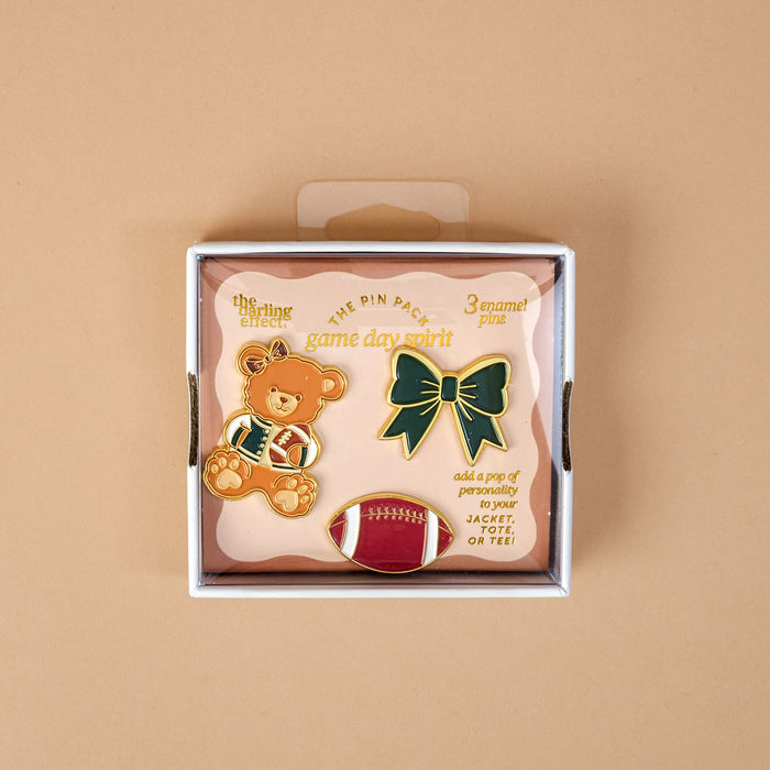 Introducing the Game Day Spirit The Pin Pack by The Darling Effect—a box featuring three expressive pins: a teddy bear in sports attire, a green bow, and a red football. The packaging reads "game day spirit" and recommends adding these collectible accessories to your jacket or tee for the perfect curated pin pack.