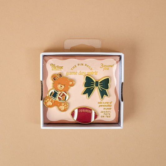 Introducing the Game Day Spirit The Pin Pack by The Darling Effect—a box featuring three expressive pins: a teddy bear in sports attire, a green bow, and a red football. The packaging reads "game day spirit" and recommends adding these collectible accessories to your jacket or tee for the perfect curated pin pack.