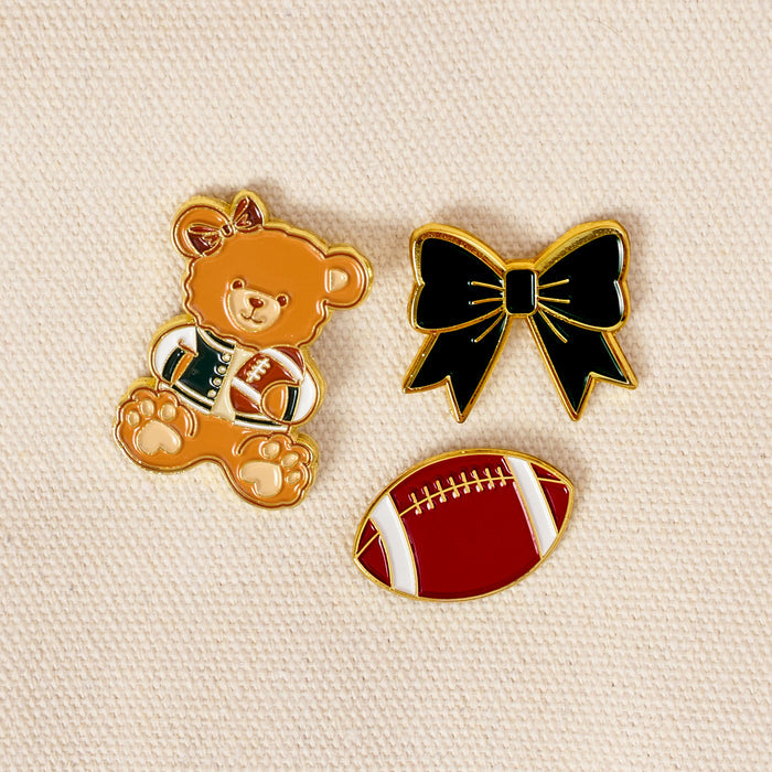 Three collectible accessories from The Darling Effect’s Game Day Spirit The Pin Pack rest on a beige textured surface: a teddy bear holding a football, a black bow with gold trim, and a red football with white stripes—an expressive pin set perfect for any curated collection.