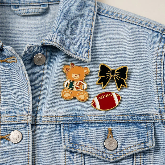 Three expressive pins from The Darling Effect’s Game Day Spirit The Pin Pack—featuring a teddy bear holding a football, a black bow, and a red football with white laces—adorn a light blue denim jacket just above the left pocket, adding unique flair to your style.