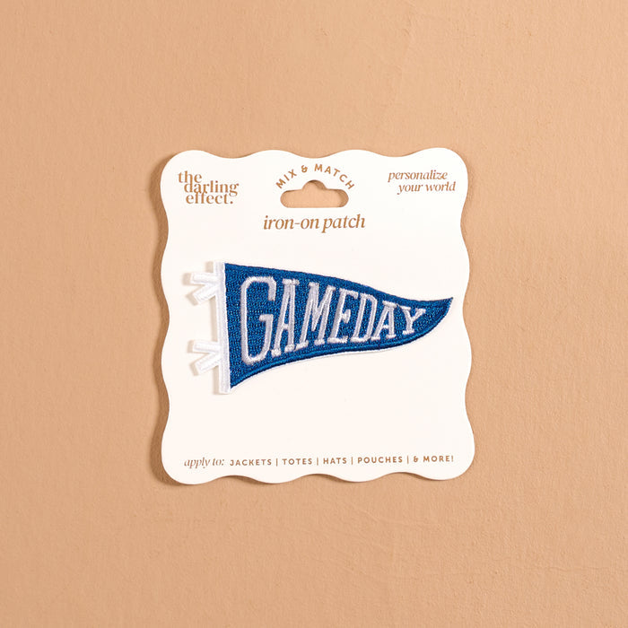A blue pennant-shaped iron-on patch reading “GAMEDAY” in white letters from The Darling Effect, the Gameday Blue Mix & Match Iron-On Patch is presented on cream packaging against a beige background. Ideal for personalizing items like jackets, totes, hats, and more, as suggested on the packaging.