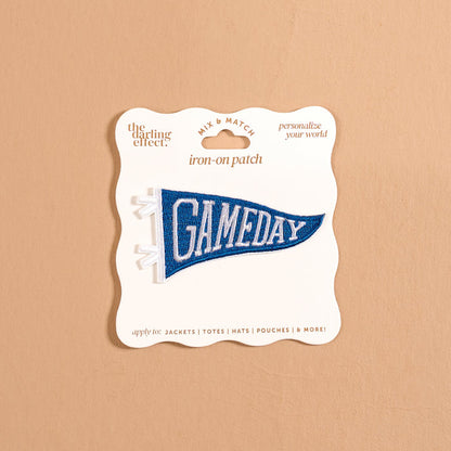 A blue pennant-shaped iron-on patch reading “GAMEDAY” in white letters from The Darling Effect, the Gameday Blue Mix & Match Iron-On Patch is presented on cream packaging against a beige background. Ideal for personalizing items like jackets, totes, hats, and more, as suggested on the packaging.