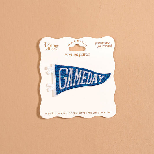 A blue pennant-shaped iron-on patch reading “GAMEDAY” in white letters from The Darling Effect, the Gameday Blue Mix & Match Iron-On Patch is presented on cream packaging against a beige background. Ideal for personalizing items like jackets, totes, hats, and more, as suggested on the packaging.