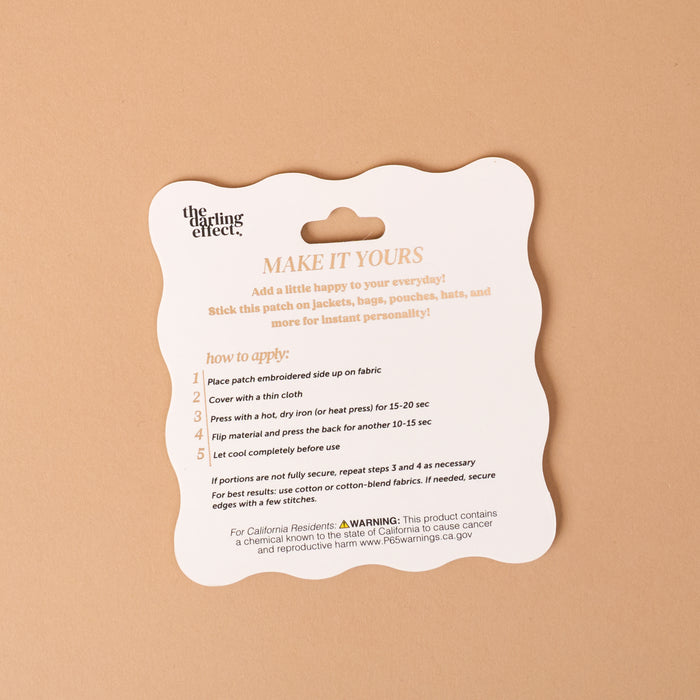 Square card on a tan background with instructions for applying The Darling Effect Gameday Blue Mix & Match Iron-On Patch. Steps include placing the patch, covering it, pressing with an iron, peeling, reheating as needed, and letting cool. Perfect for using these colorful patches to customize your favorite items. Warning and website information appear at the bottom of the card.