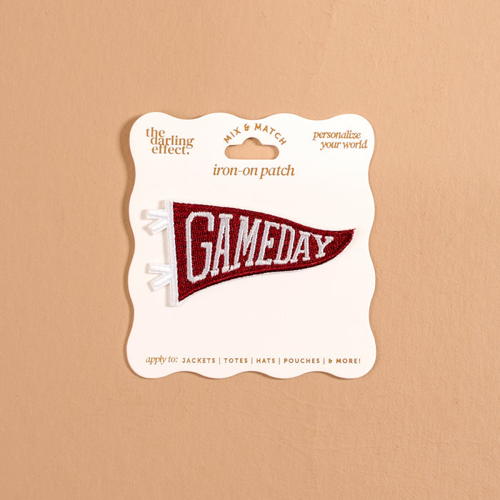 A burgundy pennant-shaped iron-on patch from The Darling Effect, featuring white "GAMEDAY" text, is attached to a cream display card against a tan background, offering a fun way to customize your gear with bold, colorful designs. This Gameday Burgundy Mix & Match Iron-On Patch is perfect for personalizing any item.