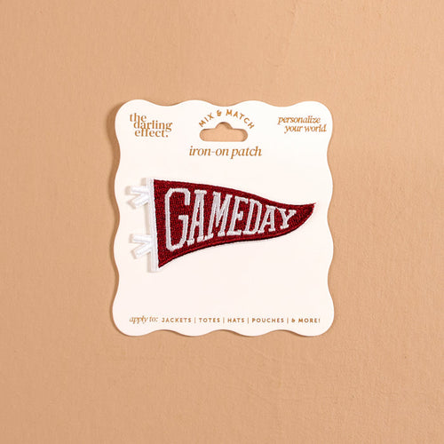 A burgundy pennant-shaped iron-on patch from The Darling Effect, featuring white "GAMEDAY" text, is attached to a cream display card against a tan background, offering a fun way to customize your gear with bold, colorful designs. This Gameday Burgundy Mix & Match Iron-On Patch is perfect for personalizing any item.
