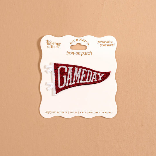 A burgundy pennant-shaped iron-on patch from The Darling Effect, featuring white "GAMEDAY" text, is attached to a cream display card against a tan background, offering a fun way to customize your gear with bold, colorful designs. This Gameday Burgundy Mix & Match Iron-On Patch is perfect for personalizing any item.
