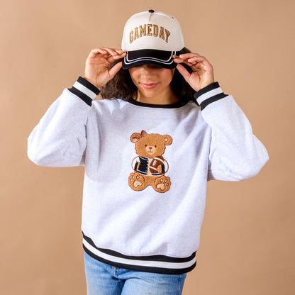 A woman wearing The Darling Effect's Gameday Classic Canvas Hat and a white sweatshirt featuring an adorable football bear graphic stands against a beige background, tilting her cap with a smile. The relaxed fit of her outfit enhances the effortless game day vibe.