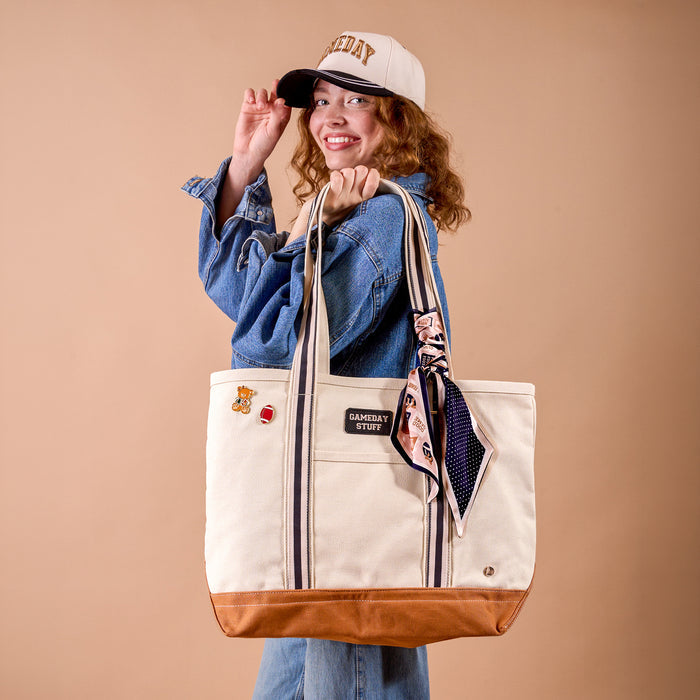 A smiling person in a denim jacket and The Darling Effect’s Gameday Classic Canvas Hat holds a large beige tote bag with brown trim, striped handles, and a scarf tied on. The bag is decorated with patches and pins. The background is a solid light brown color.