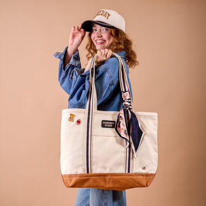 A smiling person in a denim jacket and The Darling Effect’s Gameday Classic Canvas Hat holds a large beige tote bag with brown trim, striped handles, and a scarf tied on. The bag is decorated with patches and pins. The background is a solid light brown color.