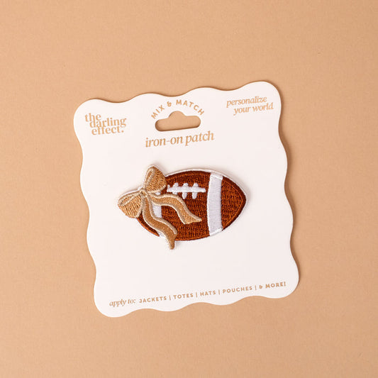 Gameday Fab Football Mix & Match Iron-On Patch by The Darling Effect features a brown football shape adorned with a gold and white bow, presented on scalloped white packaging against a tan background. Ideal for personalizing your wardrobe, this iron-on patch allows you to easily add sporty flair to any item.