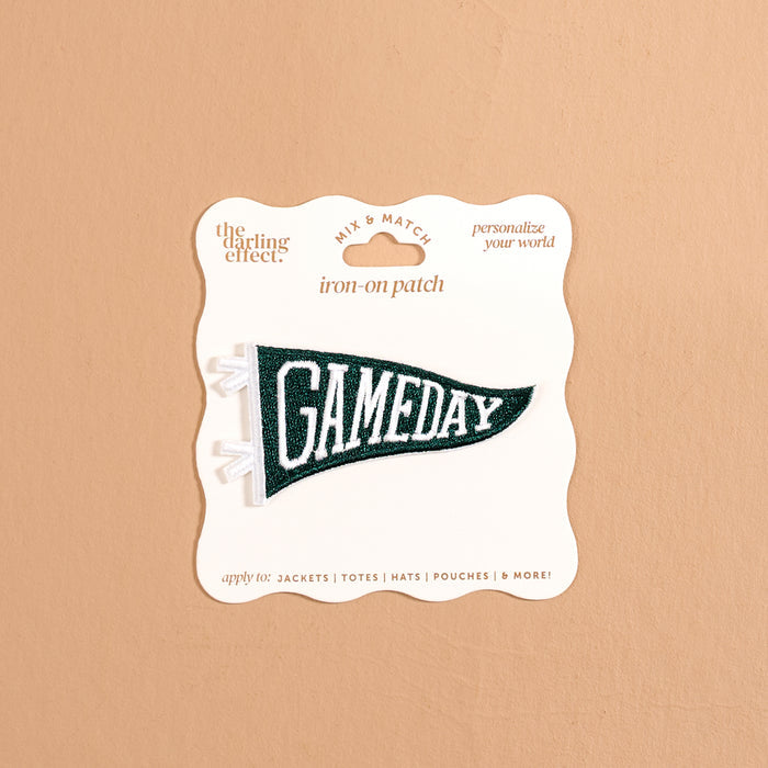 The Darling Effect’s Gameday Green Mix & Match Iron-On Patch features a green pennant shape with bold white “GAMEDAY” lettering, attached to a beige card labeled “iron-on patch” and “personalize your world.” Perfect for anyone looking to customize their gear, this patch is showcased against a neutral tan background.