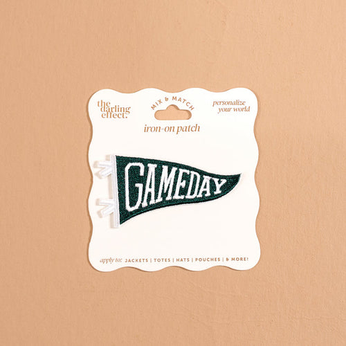 The Darling Effect’s Gameday Green Mix & Match Iron-On Patch features a green pennant shape with bold white “GAMEDAY” lettering, attached to a beige card labeled “iron-on patch” and “personalize your world.” Perfect for anyone looking to customize their gear, this patch is showcased against a neutral tan background.