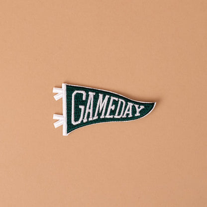 A Gameday Green Mix & Match Iron-On Patch by The Darling Effect, featuring bold green and white felt with the word GAMEDAY, is displayed on a simple beige background—perfect for customizing with colorful designs or additional patches.