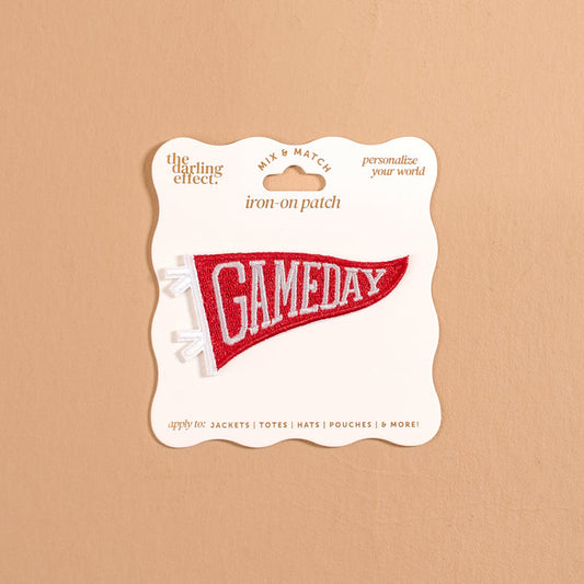 The Darling Effect's Gameday Red Mix & Match Iron-On Patch features a bold, red pennant shape with GAMEDAY in crisp white text, presented on a beige card with decorative edges against a tan background—ideal for anyone looking to personalize jackets with vibrant patches or bring extra team spirit to their iron-on patch collection.