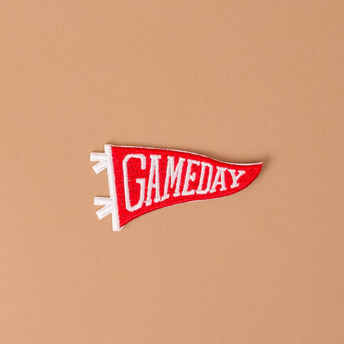 A vibrant red, pennant-shaped Gameday Red Mix & Match Iron-On Patch by The Darling Effect, featuring bold white "GAMEDAY" lettering, pops against a simple light brown background — an ideal choice for anyone wanting to customize jackets with eye-catching color or achieve instant style with easy iron-on application.