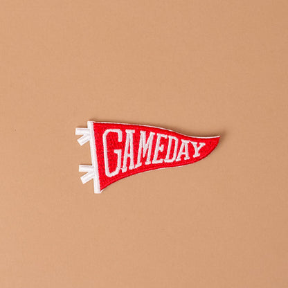 A vibrant red, pennant-shaped Gameday Red Mix & Match Iron-On Patch by The Darling Effect, featuring bold white "GAMEDAY" lettering, pops against a simple light brown background — an ideal choice for anyone wanting to customize jackets with eye-catching color or achieve instant style with easy iron-on application.
