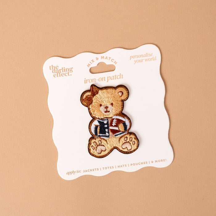 The Gameday Touchdown Teddy Mix & Match Iron-On Patch by The Darling Effect features a teddy bear in a letterman jacket, displayed on a white card with wavy edges against a beige background. Part of our colorful patch collection, this vibrant patch is perfect for adding a playful touch to jackets, totes, hats, and pouches.