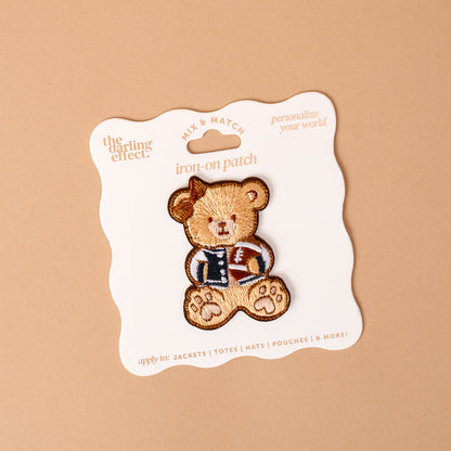 The Gameday Touchdown Teddy Mix & Match Iron-On Patch by The Darling Effect features a teddy bear in a letterman jacket, displayed on a white card with wavy edges against a beige background. Part of our colorful patch collection, this vibrant patch is perfect for adding a playful touch to jackets, totes, hats, and pouches.