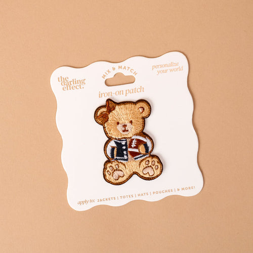 The Gameday Touchdown Teddy Mix & Match Iron-On Patch by The Darling Effect features a teddy bear in a letterman jacket, displayed on a white card with wavy edges against a beige background. Part of our colorful patch collection, this vibrant patch is perfect for adding a playful touch to jackets, totes, hats, and pouches.