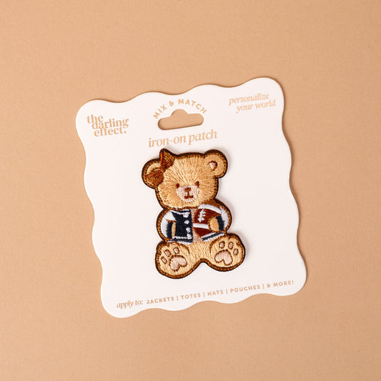 The Gameday Touchdown Teddy Mix & Match Iron-On Patch by The Darling Effect features a teddy bear in a letterman jacket, displayed on a white card with wavy edges against a beige background. Part of our colorful patch collection, this vibrant patch is perfect for adding a playful touch to jackets, totes, hats, and pouches.